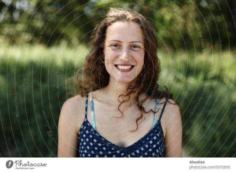 direct portrait of young beautiful brunette woman smiling full of joy and standing in front of a piece of forest warm Summery Beautiful weather plants proximity