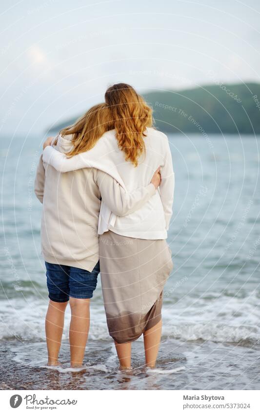 Redhead senior mother and her adult beautiful daughter are walking together on the sea shore. embrace vertical back unrecognizable seashore nature people