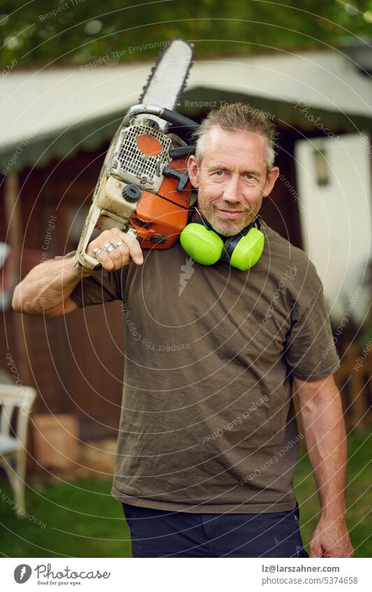 Middle-aged gardener holding a chainsaw over his shoulder after work with ear protection looking at the camera craftsman outside concept craftsmanship worker