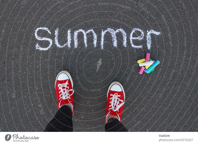 Top view of the word summer written with white chalk on the sidewalk, colored chalk and legs in red sneakers. Chalk asphalt colorful crayons childhood drawing
