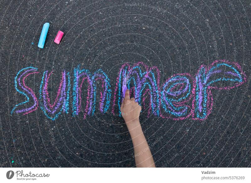 The child writes the word summer on the pavement with multi-colored chalk. Top view of the hand. Chalk asphalt colorful crayons childhood drawing street