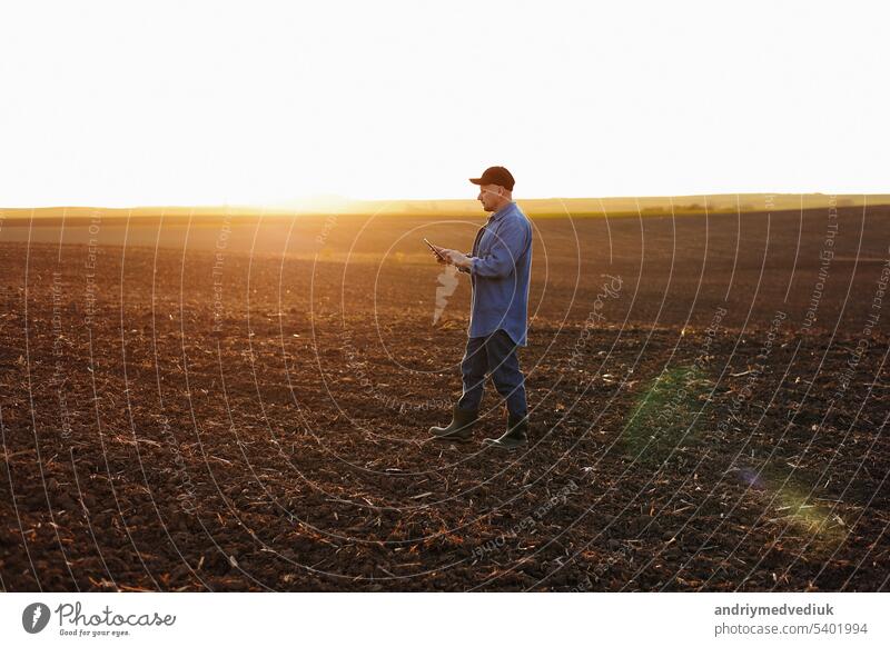 Smart farming technology and agriculture. Farmer uses digital tablet on field with plowed soil at sunset. Checking and control of soil quality, land readiness for sowing crops and planting vegetables