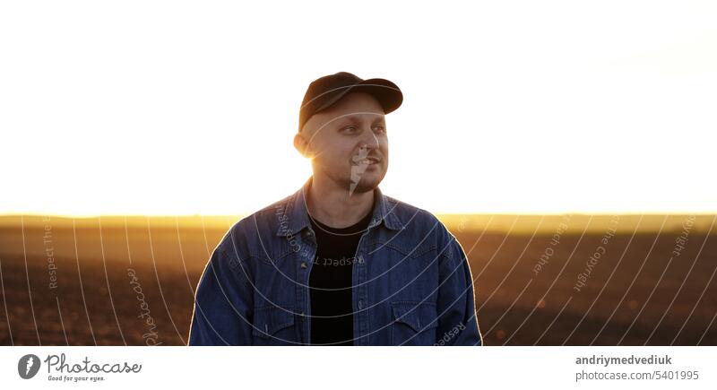 Portrait of smiling male farmer in cap is walking through endless plowed farmland field on sunset. Professional agronomist inspecting cultivated land in sunlight. Agribusiness concept and farming.