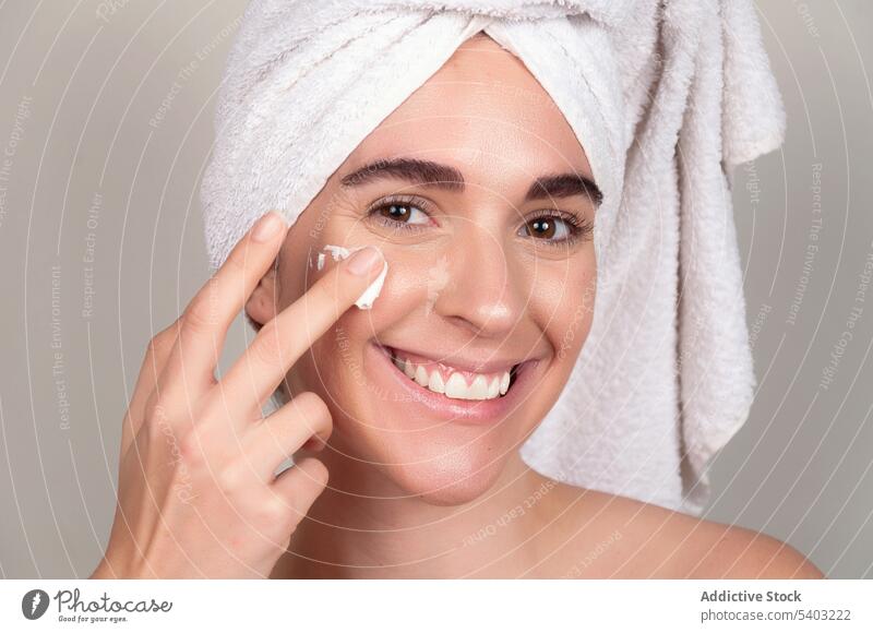 Smiling woman applying cosmetic cream on face skin care smile moisture towel treat finger bare shoulders hydrate young female beauty cosmetology procedure