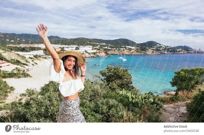 Happy woman on tropical resort looking at camera with raised arm summer vacation sea hat smile female balearic islands tourism travel seascape ocean coast shore