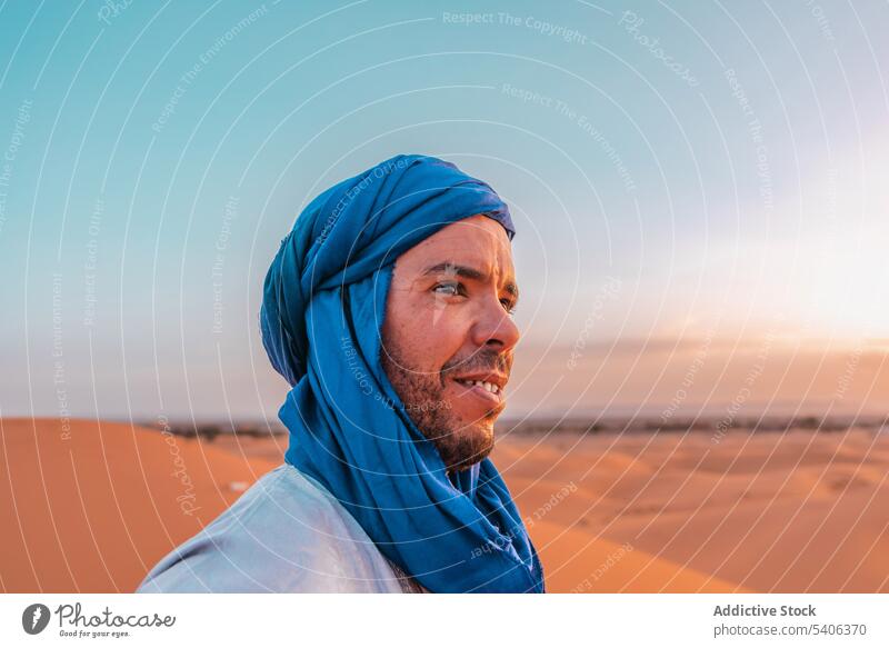 Smiling ethnic man in blue turban standing in desert berber tradition happy contemplate tuareg sand nature merzouga morocco muslim blue sky headwear adventure