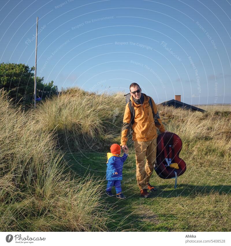 Young man with toddler and child seat leaves a hut in a dune landscape in Denmark Father Toddler Child seat Longing Look back Safety Marram grass Hut Infancy