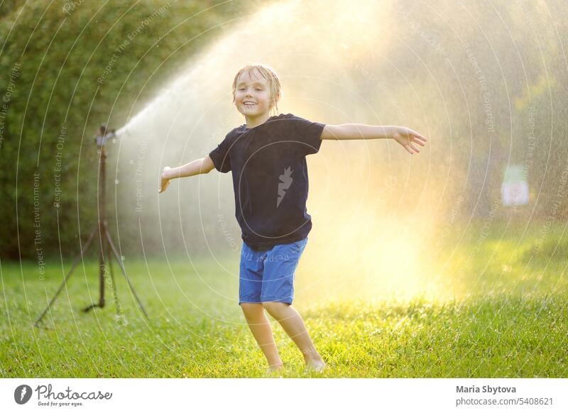 Funny little boy playing with garden sprinkler in sunny city park. Elementary school child laughing, jumping and having fun with spray of water. Summer outdoors activity for kids.