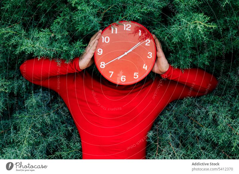 Man hiding face with red clock against green bushes man cover face hide time park nature deadline punctual male casual plant shrub garden minute daytime foliage