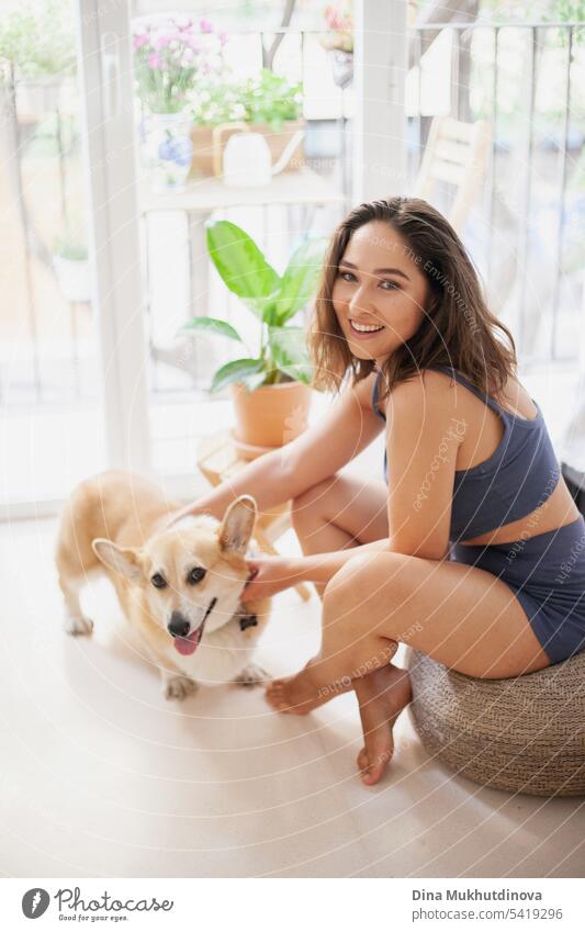 millennial caucasian woman at home with corgi dog. Funny cozy picture of female with puppy in apartment. Brunette smiling with welsh corgi Pembroke dog sitting on couch.
