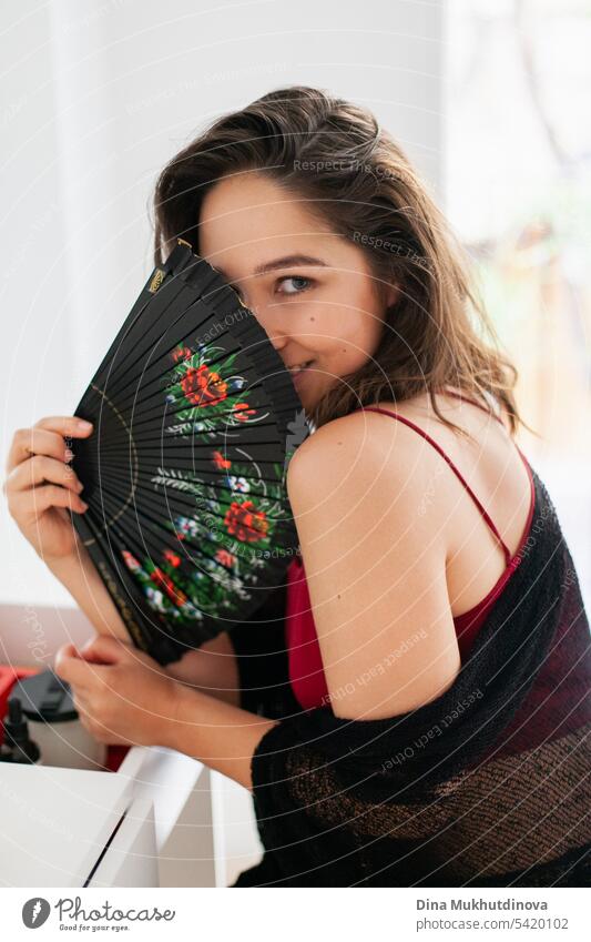 Sensual smiling woman in red and black outfit using fan. Spanish vacation. Passionate romantic female portrait in boudoir room. Summer heatwave.