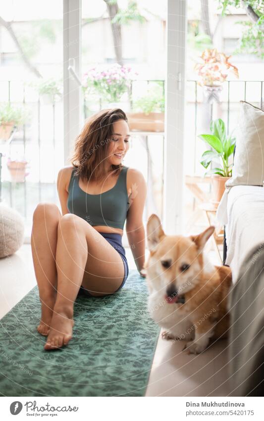 young woman with corgi Pembroke dog doing yoga on green mat at home. Practicing sport, wellness and healthcare. Indoor yoga online class. Mindfullnesss and meditation, active lifestyle.