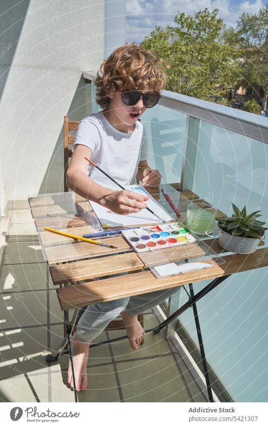 Cheerful boy painting on balcony watercolor table smile container brush art creative kid child draw childhood tool sit happy lifestyle home apartment sunny