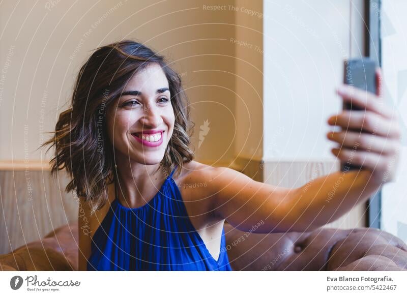 young beautiful woman taking a selfie with mobile phone and smiling. wearing a casual blue dress. Indoors, technology and lifestyle portrait person camera
