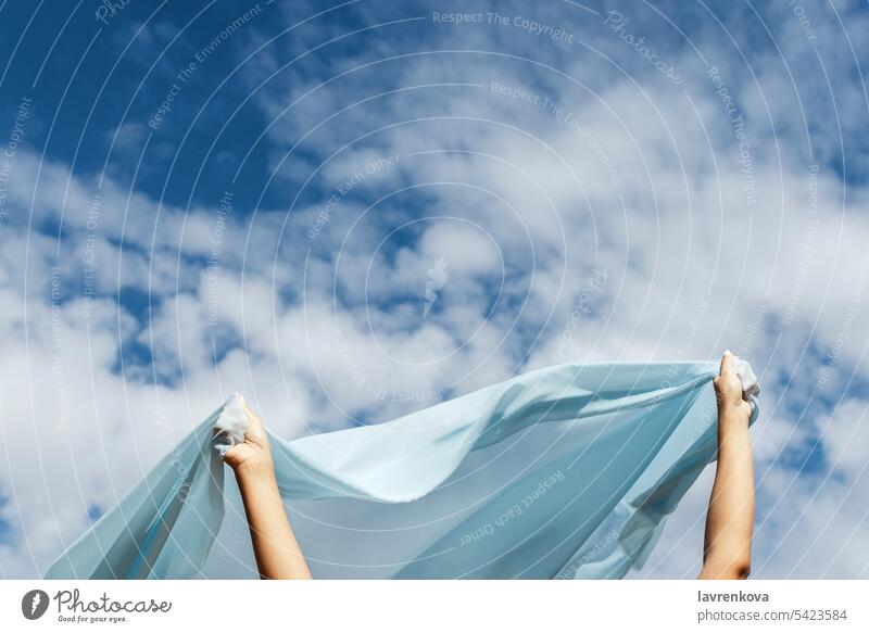 Female hands holding cut of blue cloth up in the sky, selective focus horizon high flight fly view air outdoor summer nature alone clouds fabric female freedom
