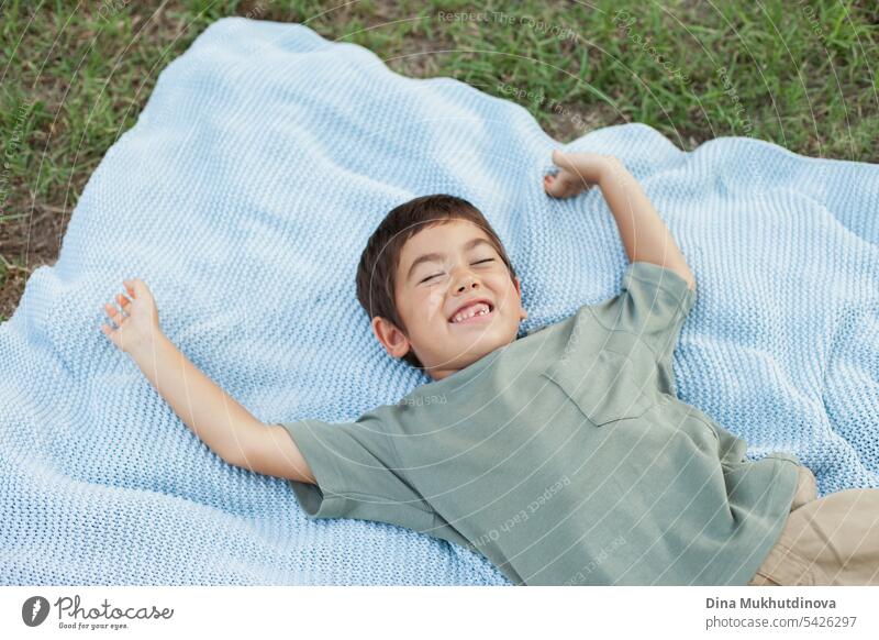 diverse kid in the park lying on the blue blanket and laughing on green grass, smiling with eyes closed. Five year old boy in the park in summer, feeling happy.
