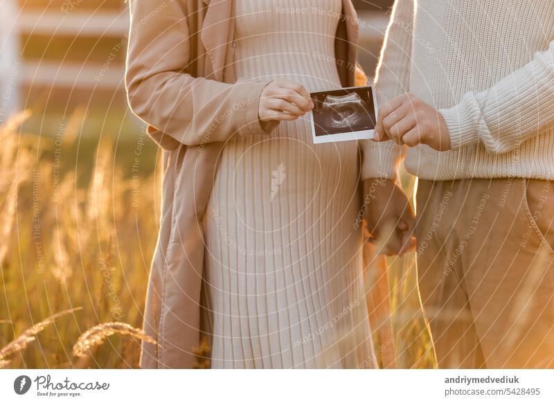 Unrecognised of a pregnant woman with her husband are holding an ultrasound scan photo of the unborn child outdoors on nature in sunlights. Happy motherhood and parenthood concept