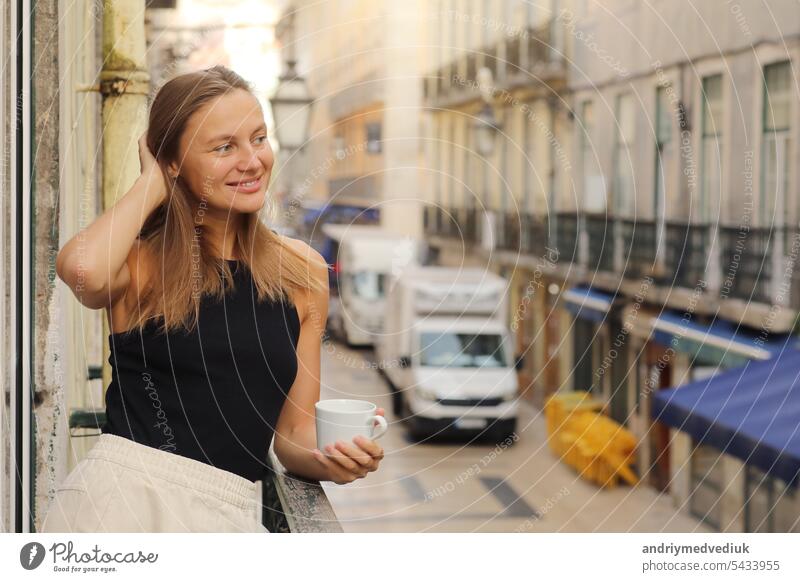 Happy young beautiful woman holds cup of coffee or tea, standing on the balcony with Europe city street in the morning. Mature female enjoying vacation in resort. Summer, weekend, tourism, travel