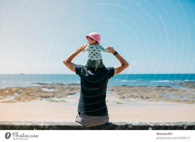 Back view of father with little daughter on his shoulders on the beach daughters pa fathers daddy dads papa beaches child children family families people