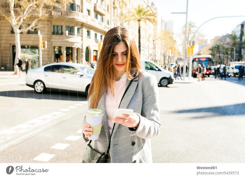 Spain, Barcelona, young woman with coffee to go standing at roadside looking at cell phone females women Coffee Smartphone iPhone Smartphones Coffee to Go