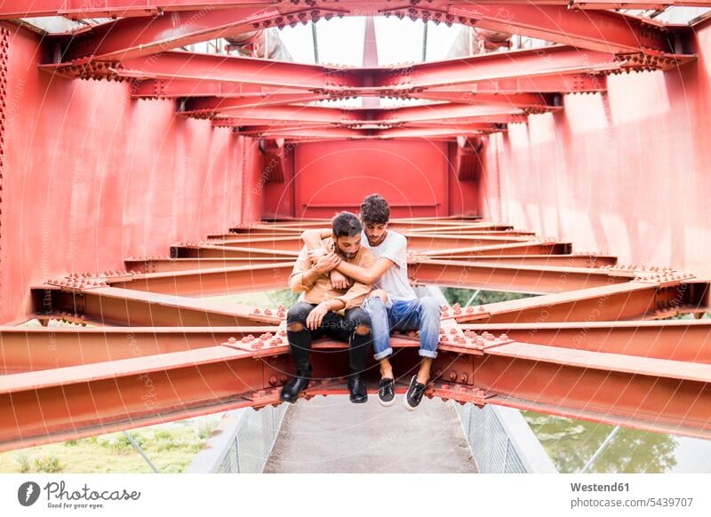 Young gay couple in love sitting on steel girder of a footbridge looking down twosomes partnership couples foot bridges Steel Girders gay men gay man