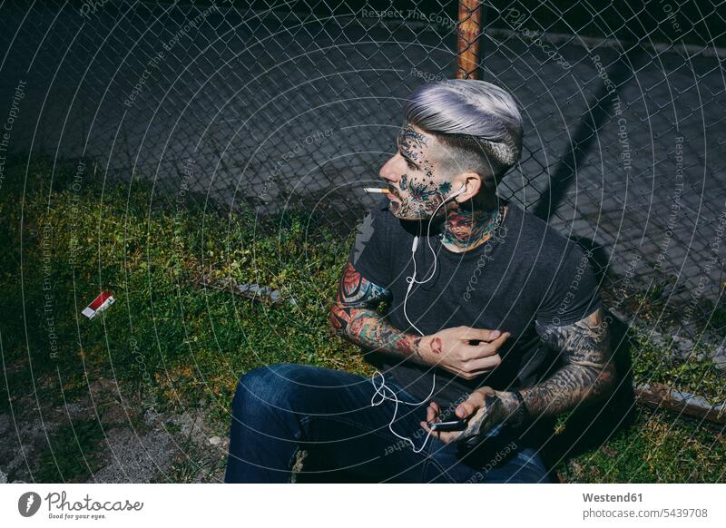 Tattooed young man with earbuds and smartphone smoking a cigarette at wire mesh fence tattooed tattoos men males Smartphone iPhone Smartphones Wire Mesh Fence