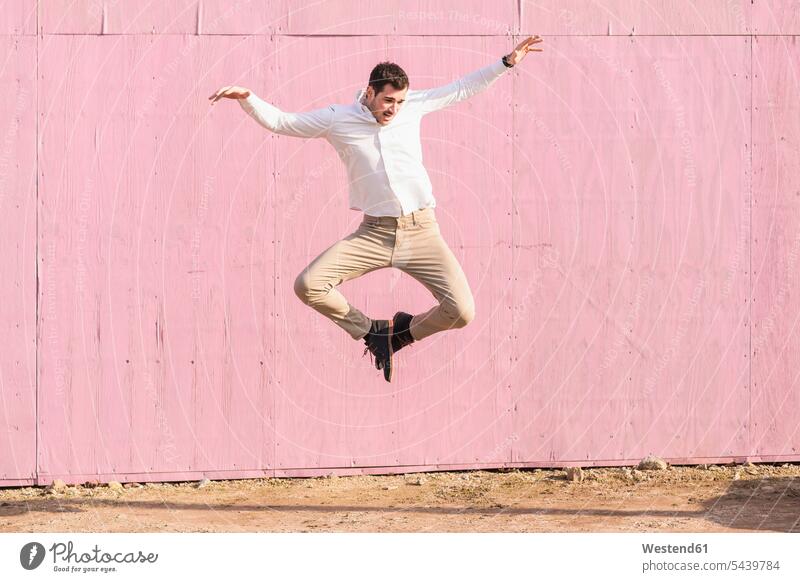 Exuberant young man jumping in front of pink wall Leaping men males walls Rosy exuberance hilarity Frolic exuberant jumps Adults grown-ups grownups adult people
