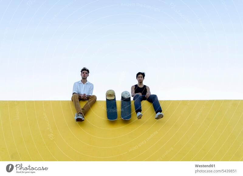 Two young men with skateboards sitting on a high yellow wall friends mate man males walls resting Skate Board Seated friendship Adults grown-ups grownups adult