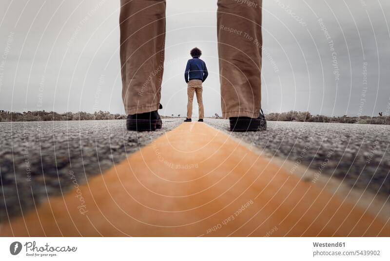 View through standing legs to a curly man standing on the medial strip of a road caucasian caucasian ethnicity caucasian appearance european Surface Level