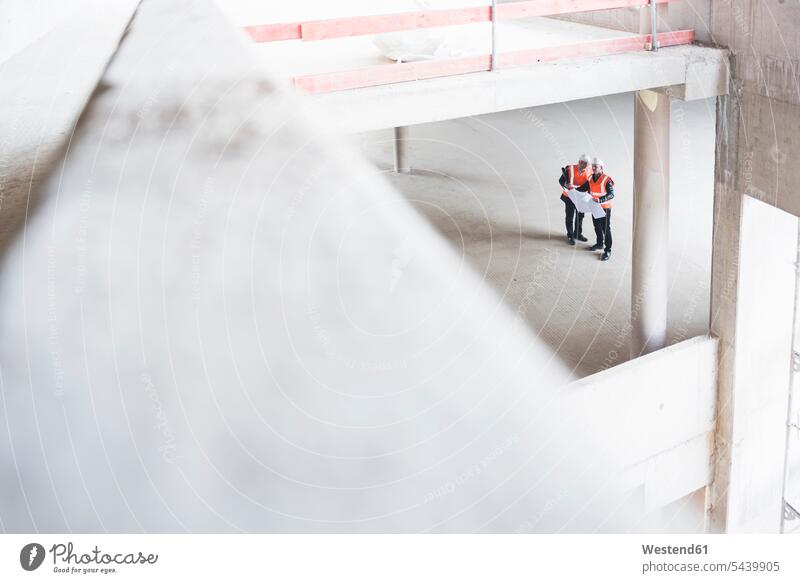 Two men with plan wearing safety vests talking in building under construction speaking working At Work man males construction site Building Site sites