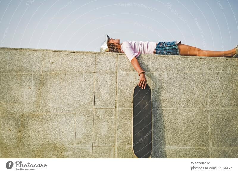 Girl with skateboard lying on wall laying down lie lying down Taking a Break resting break baseball cap visor cap peaked cap visored cap leisure free time