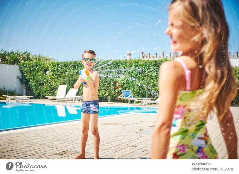 Boy with water gun splashing at girl at the poolside pool edge Pool Side swimming pool swimming pools females girls boy boys Water gun Water guns squirt gun