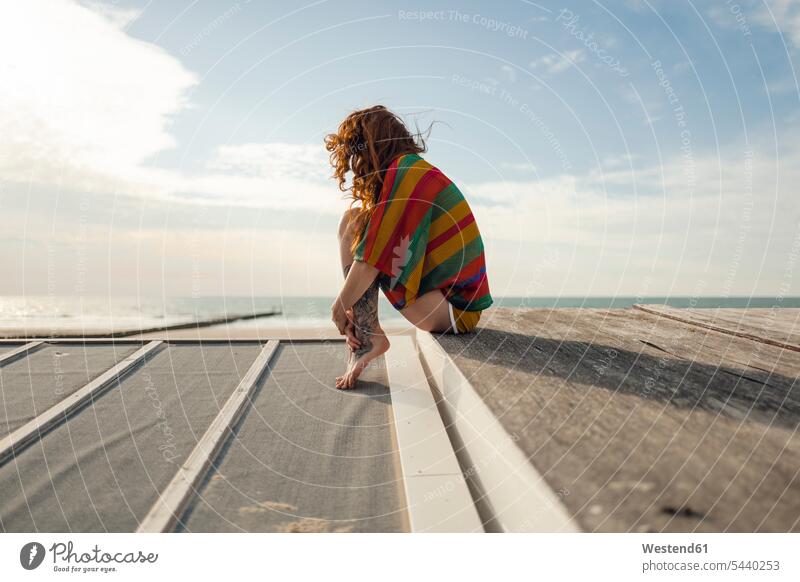 Woman with tattoo crouching on plank, looking at the sea beach beaches ocean planks woman females women perching Perched sitting Seated water waters