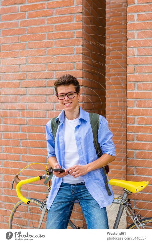 Portrait of smiling young man with racing cycle and cell phone standing in front of brick wall men males portrait portraits Adults grown-ups grownups adult