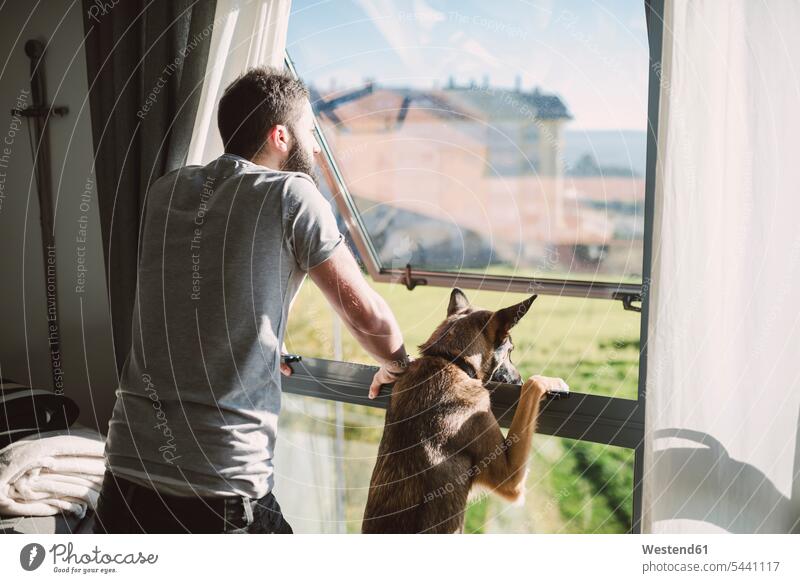 Young man standing at window with his dog, waiting young dogs Canine home at home windows men males pets animal creatures animals Adults grown-ups grownups