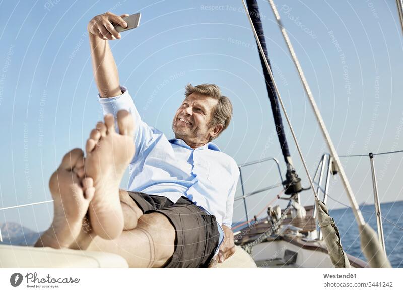 Smiling mature man lying on deck of his sailing boat taking selfie with cell phone Selfie Selfies men males boat sports Adults grown-ups grownups adult people