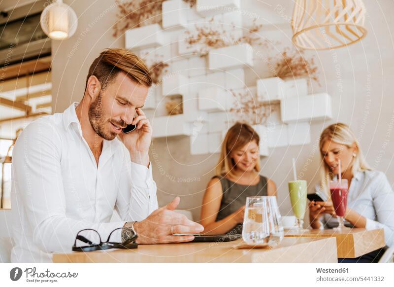 Man on cell phone in a cafe with two women in background Businessman Business man Businessmen Business men males mobile phone mobiles mobile phones Cellphone