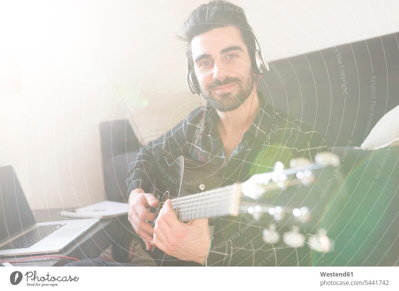 Young man at home playing guitar and wearing headphones connected to laptop smiling smile guitars Laptop Computers laptops notebook men males