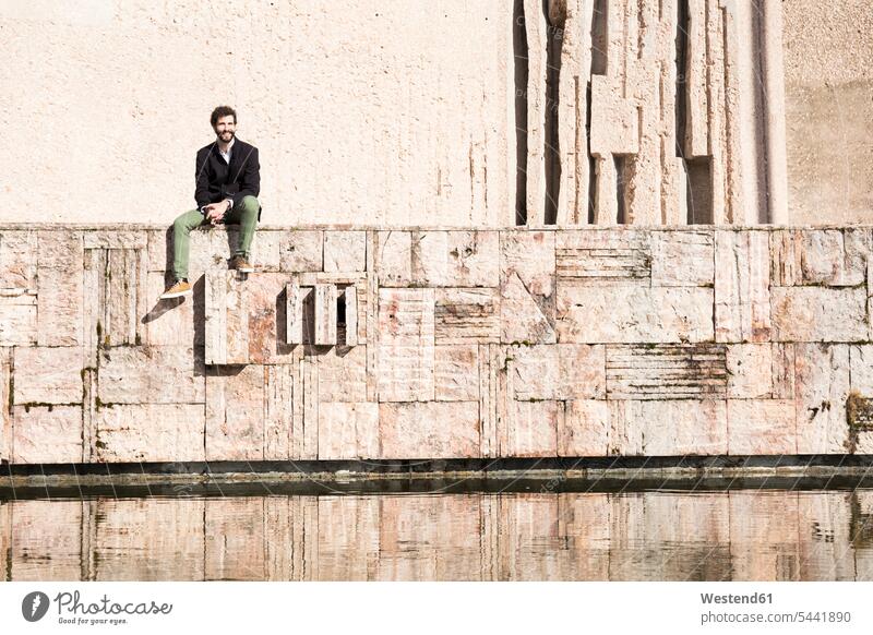 Smiling young man relaxing near water men males Adults grown-ups grownups adult people persons human being humans human beings wall walls sitting Seated