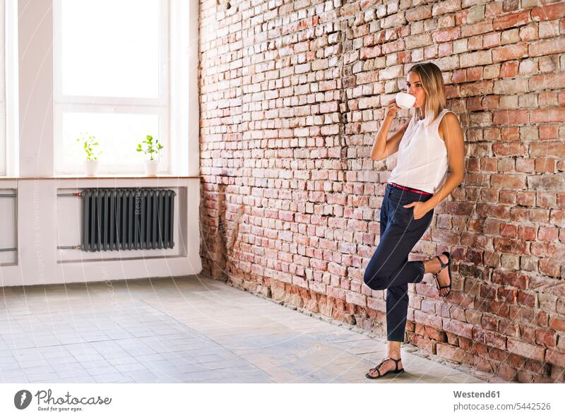 Businesswoman drinking cup of coffee at brick wall in office businesswoman businesswomen business woman business women Coffee females business people