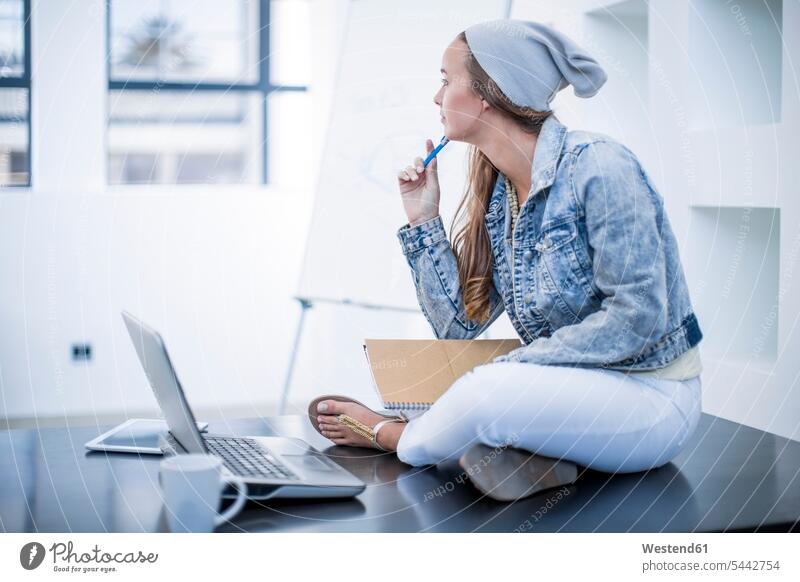 Young woman working in office sitting cross-legged on desk with her laptop desks Seated Laptop Computers laptops notebook happiness happy office worker Joy