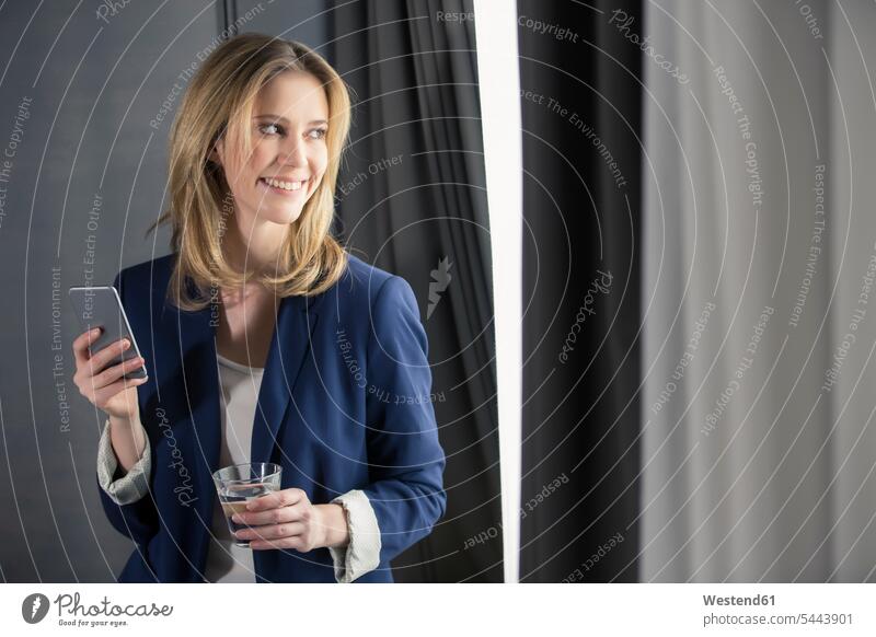 Smiling businesswoman holding glass of water and cell phone at the window businesswomen business woman business women smiling smile mobile phone mobiles