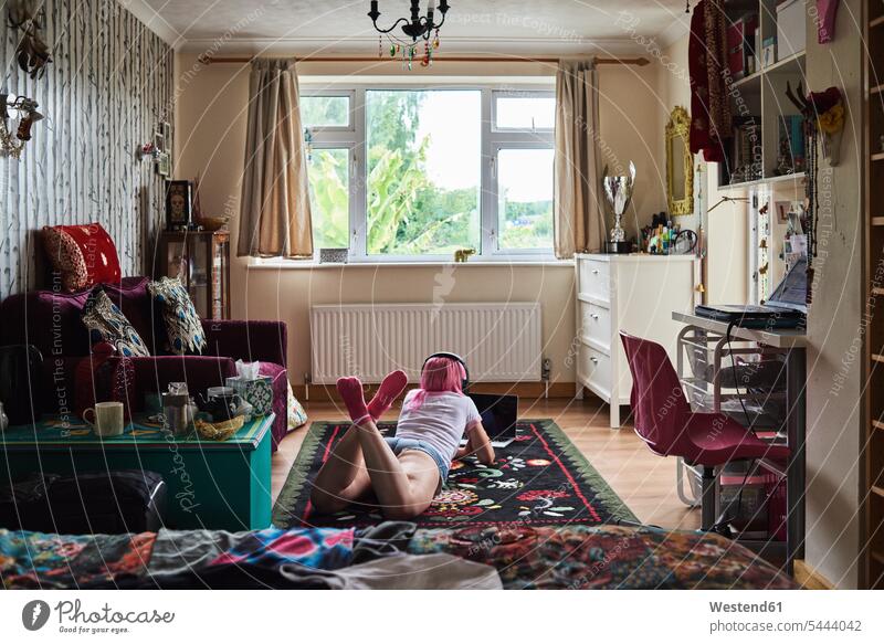 Young woman with pink hair lying on carpet wearing headphones and using laptop at home headset females women Laptop Computers laptops notebook laying down lie