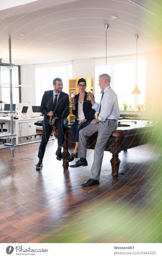 Colleagues in office sitting on pool table, toasting with bottles businesswear business attire business clothing clinking cheers work-life balance