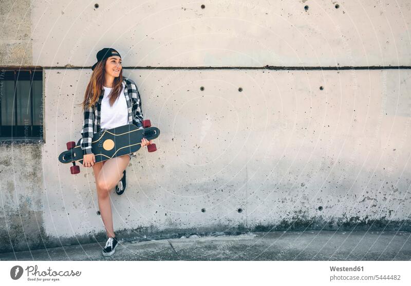 Portrait of happy young woman with longboard standing in front of concrete wall females women Adults grown-ups grownups adult people persons human being humans