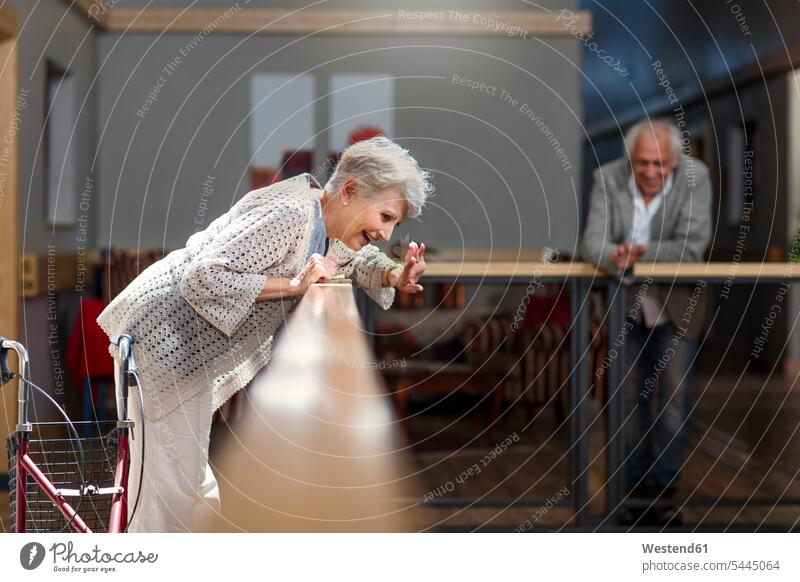 Seniors looking down from railing, woman waving and laughing standing retirement home nursing home watching observing observe wave senior women elder women