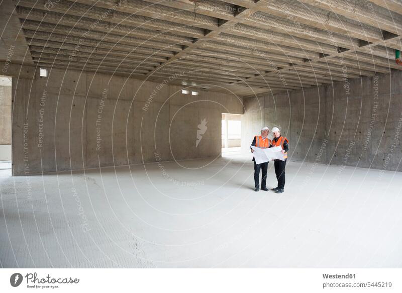 Two men with plan wearing safety vests talking in building under construction speaking working At Work construction site Building Site sites Building Sites