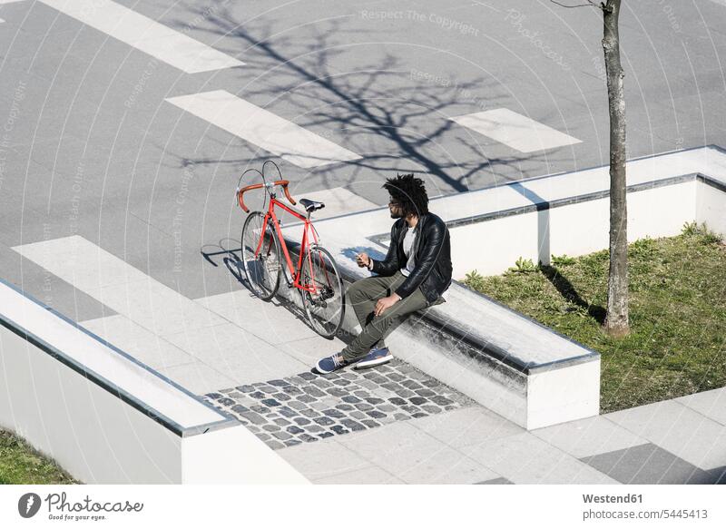 Man sitting in city skatepark holding his smartphone next to his bicycle bikes bicycles Seated man men males Adults grown-ups grownups adult people persons