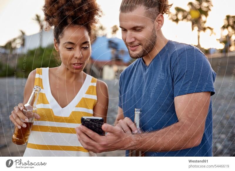 Two friends with beer bottles looking at cell phone on the beach beaches portrait portraits friendship eyeing evening in the evening watching Smartphone iPhone