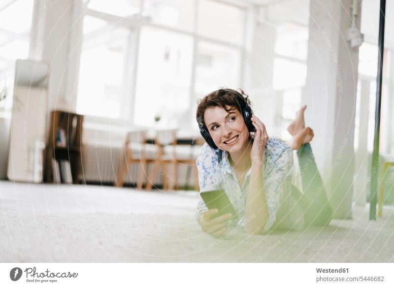 Woman lying on floor in her apartment, using smartphone and headphones hearing woman females women headset music Floor Floors Smartphone iPhone Smartphones